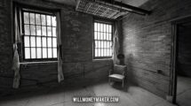 Black-and-white photograph of an abandoned room with tall windows, a chair, and a fan.