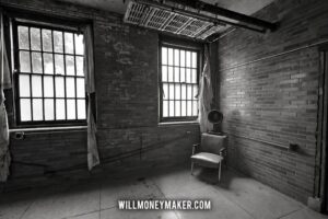 Black-and-white photograph of an abandoned room with tall windows, a chair, and a fan.