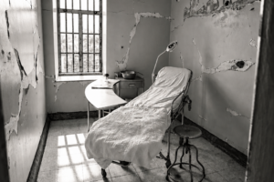 Black-and-white photograph of an empty chair beside a window in a worn room.